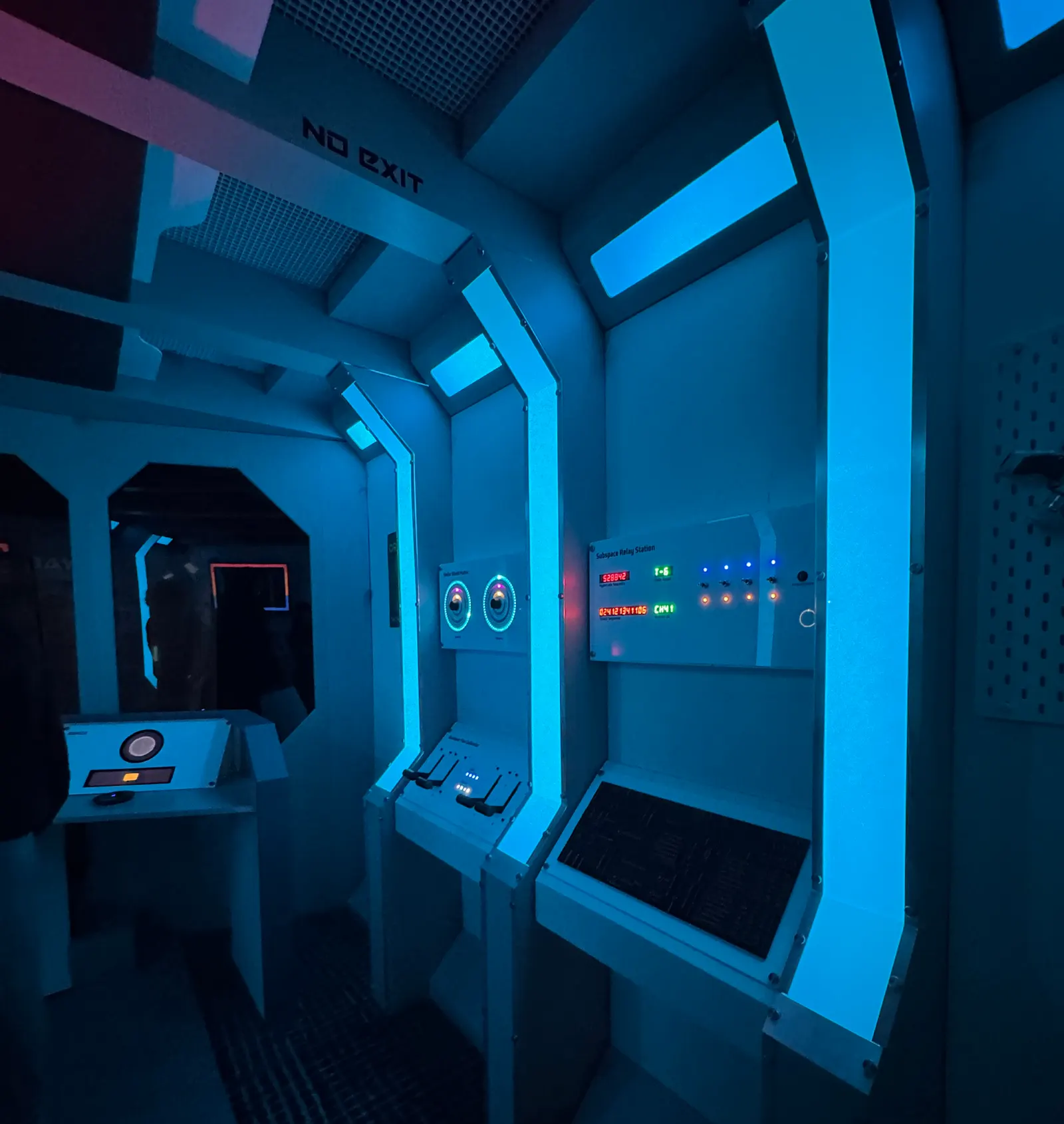 Dimly lit escape-room corridor with blue neon panels, labeled Subspace Relay Station controls, and dials