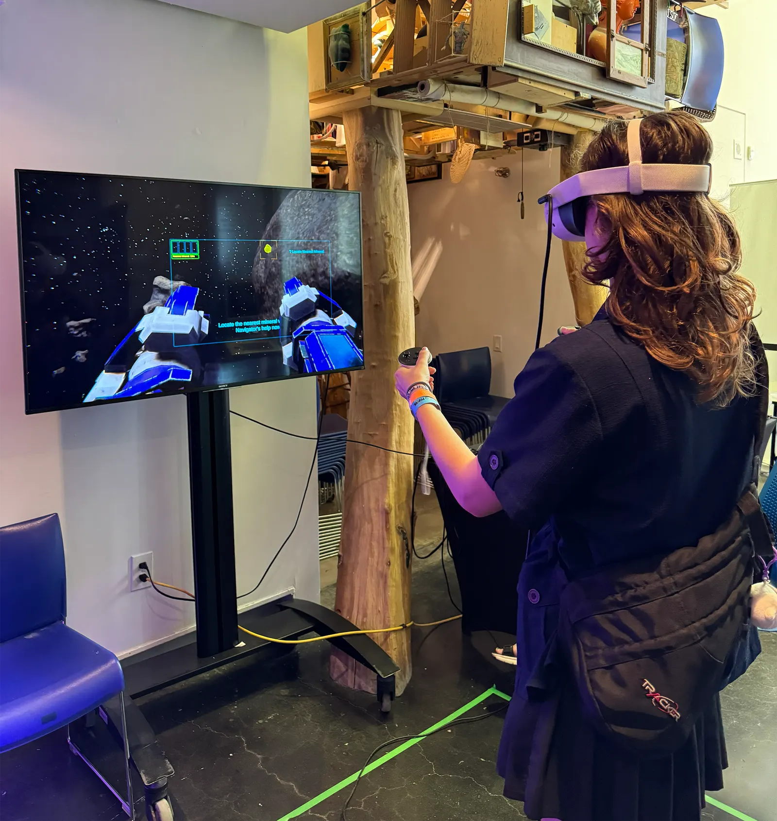 A participant in a VR headset faces a large monitor mirroring a first-person space scene with asteroid debris