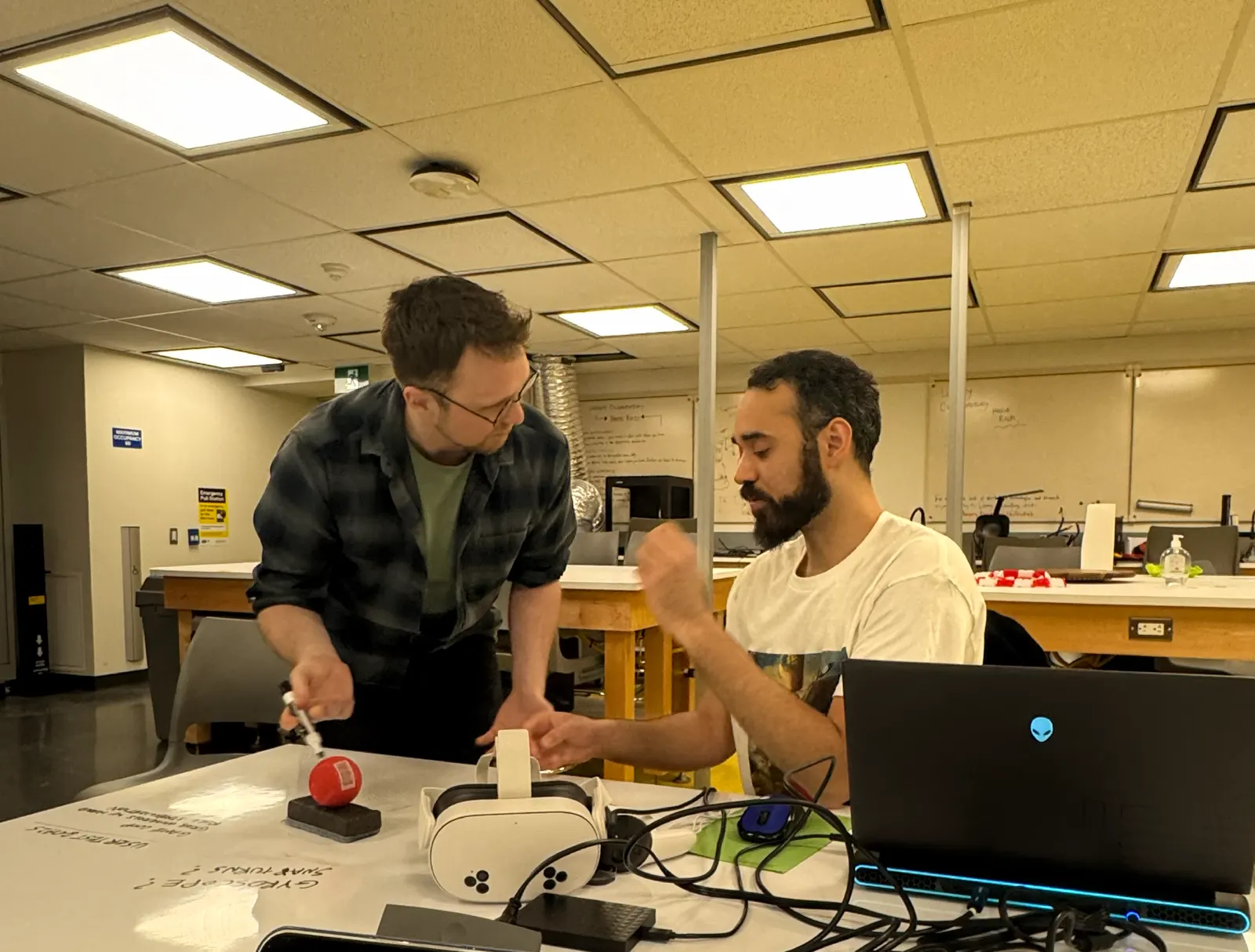 Two collaborators discuss hardware at a lab table with a VR headset, laptop, cables, and notes written on the table surface