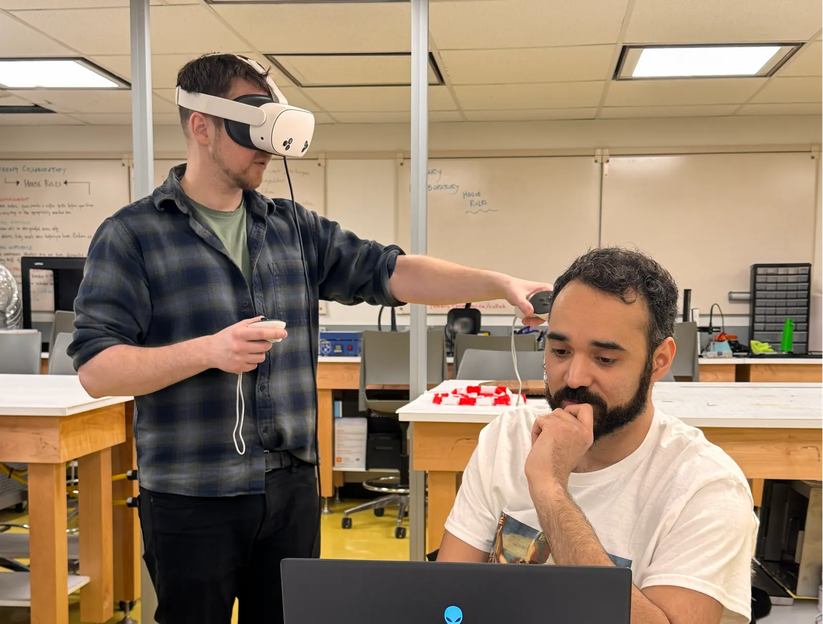 One collaborator wears a VR headset while another reviews a laptop in a workshop development space